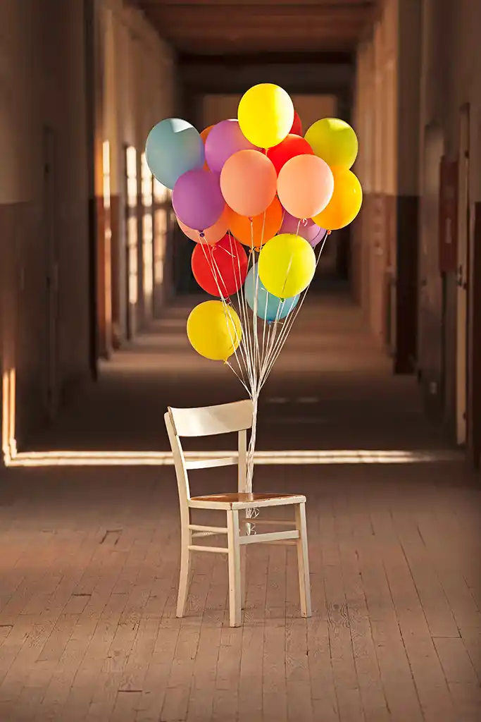 eine ganze Traube von verschiedene, farbigen Ballonen in den Farben gelb, Lila, orange, rosa hellblau und grün ist an einen stuhl gebunden, der auf einem sonst leeren, sehr langen flur steht. das bild vermittelt durch die Vielzahl an farben eine heitere fröhlichkeit