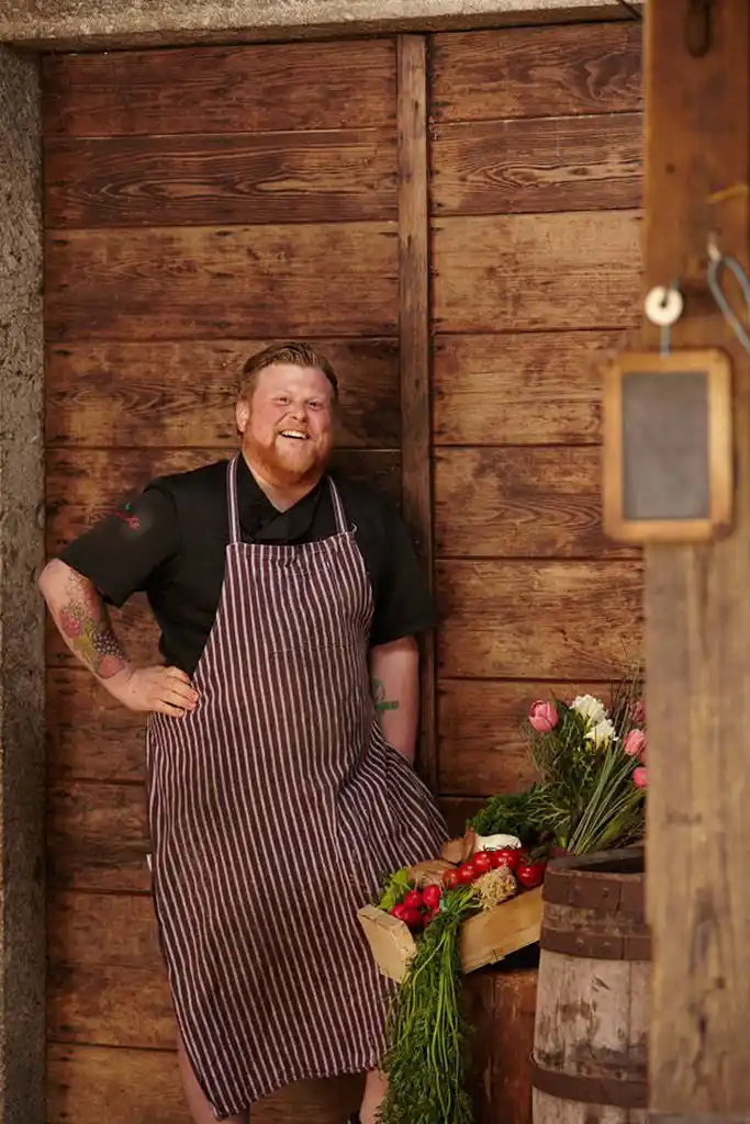 Der Koch Timo Franke steht mit einer Steige voller Gemüse vor der Tür der Scheune seiner Grosseltern und lächelt herzlich  in die Kamera.,