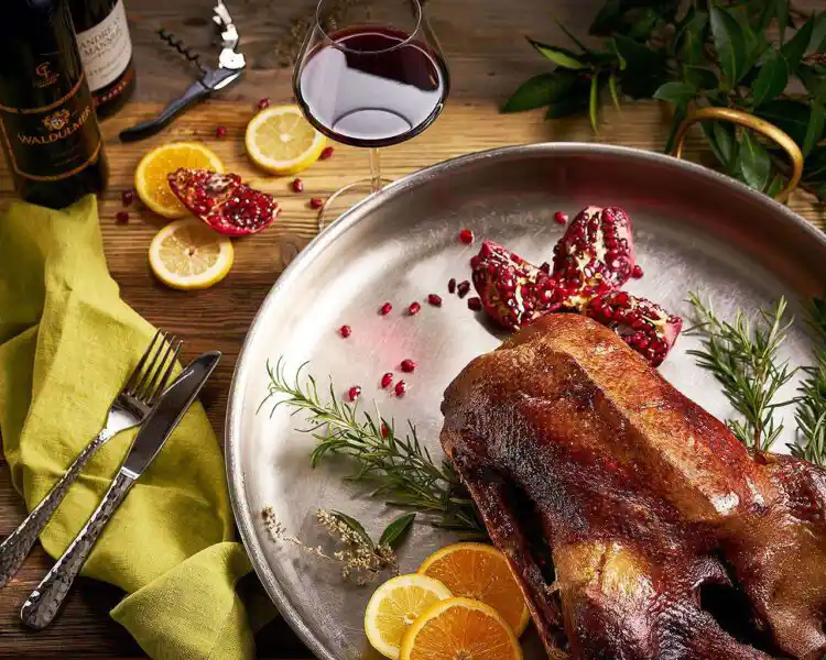 in einer großen Ofenpfanne liegt eine knusprig gebrateten Gans, der Tisch ist dekoriert mit Orangene, Glas Rotwein, einer Flasche, Rosmarin und einer Serviette. Das sieht alles sehr appetitlich aus und macht Lust auf ein Gänsebratenessen.