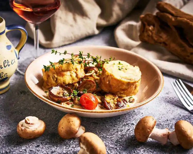 auf einem teller angerichtet liegen in scheiben geschnittene Serviettenknödel mit einer Rahmsauce mit Champignons. Vor der Teller liegen Pilze zur Dekoration, ein rustikaler Weinkrug ist am Bildrand angeschnitten und eine Gabel ragt ins Bild.Das Licht streift über das Gericht und lässt es sehr frisch und lecker aussehen. 