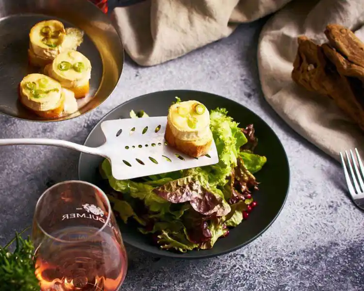 In einem schwarzen Teller ist frisch glänzender Salat angerichtet, der mit einigen roten Granatapfelkernen belegt ist. Über dem Salat schwebt eine Scheibe Baguette, auf der ein überbackener Ziegenkäse mit geschmolzener Schicht den Eindruck erweckt, er kommt gerade aus dem Ofen. Ein Gals Rosewein regt den Appetit an, ebenso wie die drei weiteren Baguette mit Schafskäse, die Links auf einem Telle ins Bild hineinragen