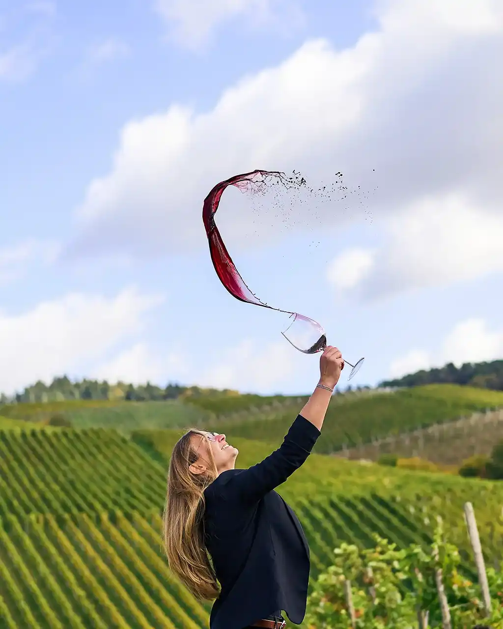 eine junge Frau mit langen haaren schleudert ein weniglasin der hand so schnell das der wein in einergeschwungenen Fontäne weit über ihren kopf schwappt