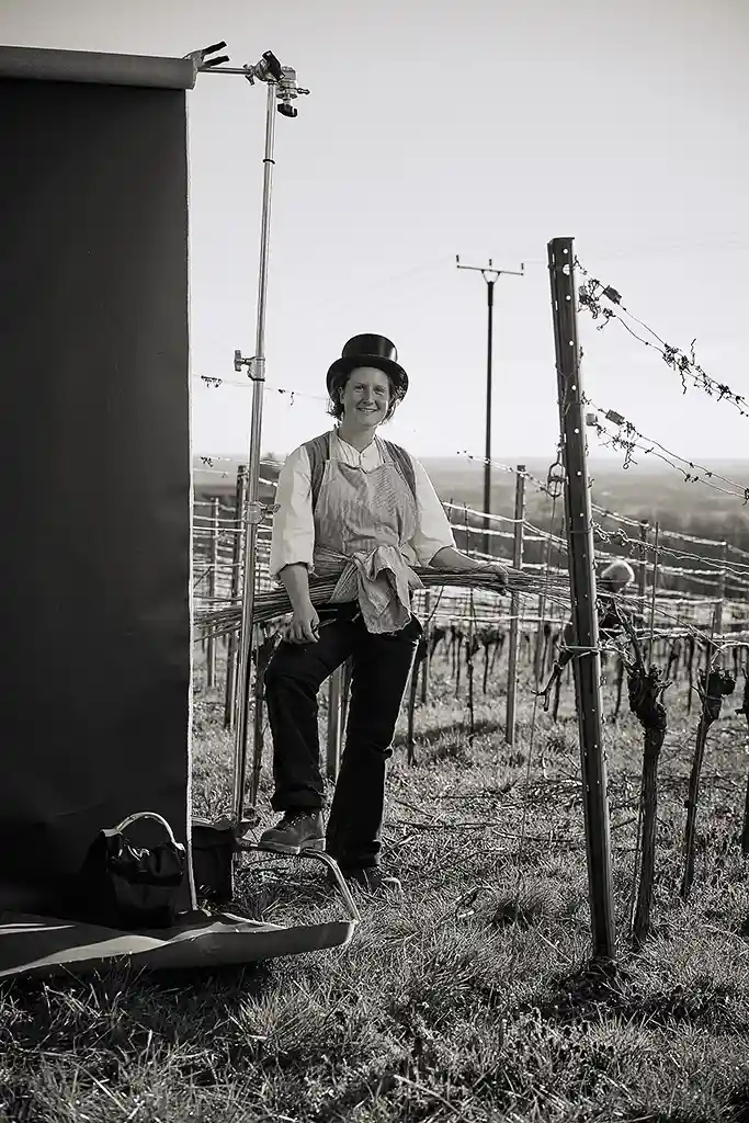 portrait der winzerin franziska im ersten jahr der walz im weinberg mit zylinder und schürze, in der sie ein buendelweidenruten trägt, mit den die Reben im Frühjahr gebunden werden